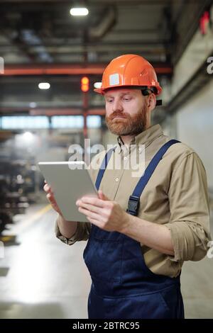 Busy young factory worker in overalls checking work file on tablet while working in industry Stock Photo