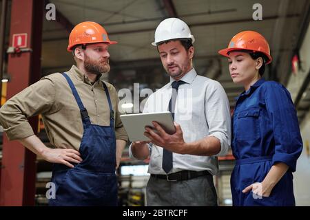 Industrial engineer in formalwear and hardhat discussing machine part with workers using 3D sketch on tablet Stock Photo