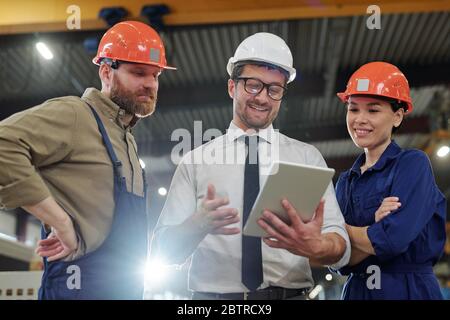 Satisfied foreman in eyeglasses watching production report with engineers while planning further work together Stock Photo