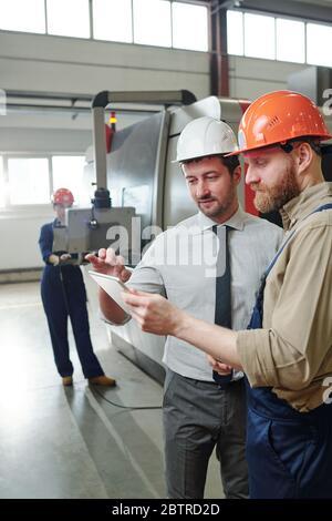 Engineers in hardhats standing in industrial shop with CNC machine and discussing online blueprint in 3D using tablet Stock Photo