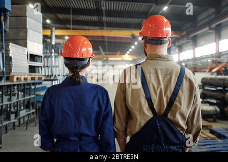 Rear view of industrial workers in orange hardhats contemplating large factory warehouse Stock Photo