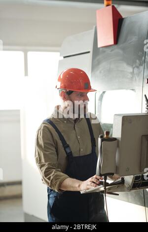 Concentrated bearded operator in hardhat and overalls choosing setups on CNC machine computer while working in factory shop Stock Photo