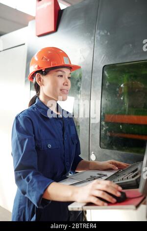 Smiling young Asian woman in hardhat operating CNC machine while checking setups on computer in factory shop Stock Photo