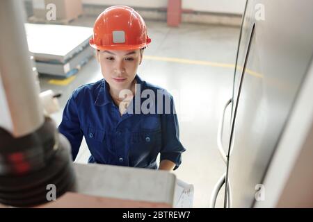 Busy Asian CNC operator in orange hardhat working on control panel of automated machine at factory Stock Photo