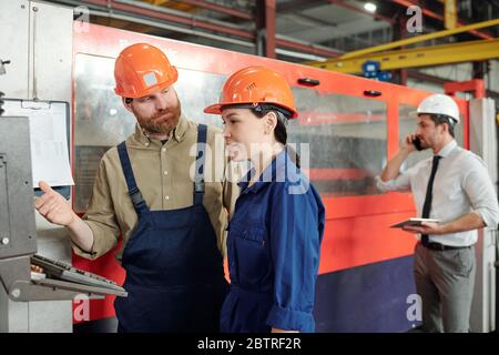 Bearded operator in hardhat teaching Asian trainee to use CNC machine in modern factory shop Stock Photo