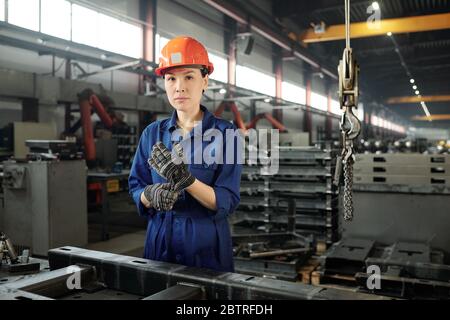 Portrait of serious young Asian woman putting safety gloves on hands in metalworking workshop Stock Photo