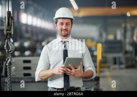 Portrait of confident handsome technical engineer in hardhat using tablet in industrial shop of large plant Stock Photo