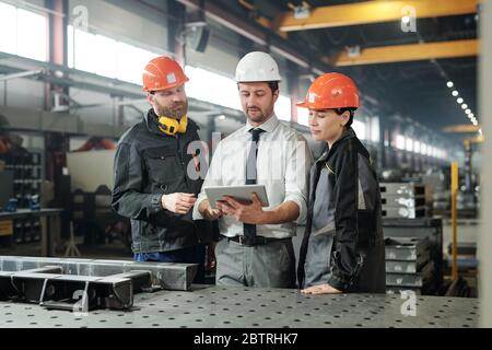 Group of engineers in hardhats using digital tablet while comparing technical drawing with finished detail at factory Stock Photo