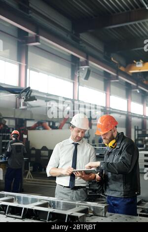Technician in formalwear using tablet while engineering industrial machine with factory worker in workshop Stock Photo