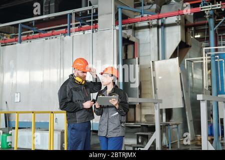 Asian woman in hardhat using digital tablet and discussing technical task with coworker in modern workshop Stock Photo