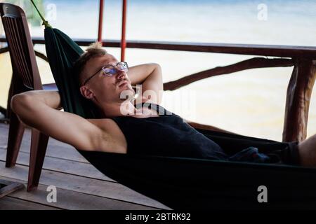 Young goodlooking man lying in a hammock in local seafood restaurant in  fishermans village Ham Ninh, tropical Phu Quoc island (Bai Thom area) in Viet Stock Photo