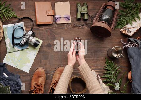 Unrecognizable hiker in sweater holding open compass above wooden table with hiking stuff and green twigs Stock Photo