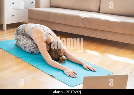 Young attractive women doing yoga exercise at home, Balasana, Child Pose, Ardha-Kurmasana Half Tortoise Pose in livingroom. Working out wearing sportswear bra and pants. Healthcare concept Stock Photo