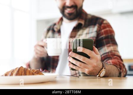 Cropped portrait of joyful bearded man using cellphone and drinking coffee while having breakfast at home Stock Photo