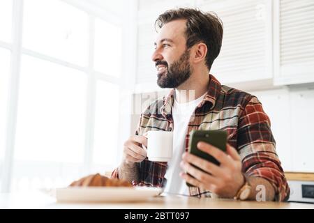 Portrait of smiling bearded man using cellphone and drinking coffee while having breakfast at home Stock Photo