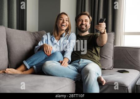 Photo of joyful beautiful nice couple watching TV and using remote control while sitting on sofa at home Stock Photo