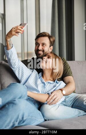 Photo of joyful beautiful nice couple smiling and taking selfie photo on cellphone while sitting on sofa at home Stock Photo