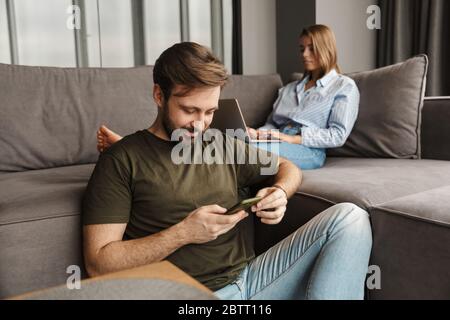 Photo of young caucasian focused couple using laptop and mobile phone while sitting at living room Stock Photo