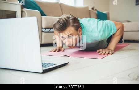 Caucasian man doing pushups on a sport carpet while looking at the laptop on the floor Stock Photo