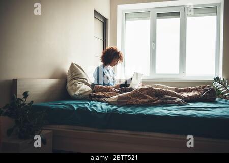 Caucasian woman with curly hair reading a book in her bed sitting near the window with a computer Stock Photo