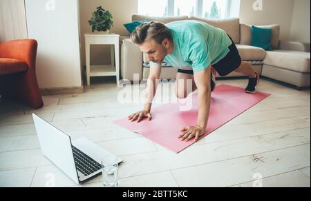 Concentrated blonde caucasian man doing stretching exercises on the yoga carpet while look at the laptop with a water glass nearby Stock Photo