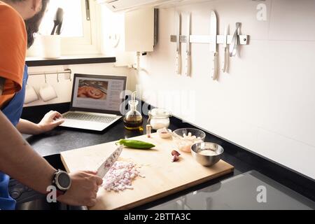 unrecognizable man consulting a recipe on the computer while chopping onions on the cutting board in the kitchen at home, concept of healthy food and Stock Photo