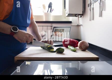 unrecognizable man cutting vegetables in the kitchen at home, in the background there is a computer with the recipe he is making, concept of healthy f Stock Photo