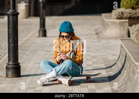 Young stylish caucasian woman skateboarder in a hat, sunglasses and jeans is using a phone next to a skate board in the city. Theme of active youth Stock Photo