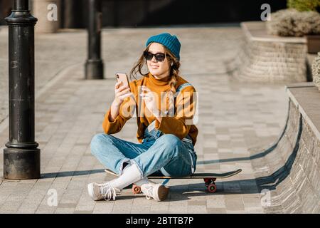 Young stylish caucasian woman skateboarder in a hat, sunglasses and jeans is using a phone next to a skate board in the city. Theme of active youth Stock Photo