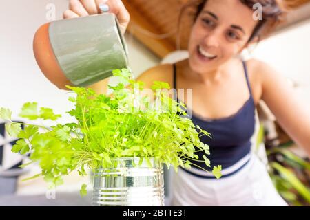 A happy caucasian woman in pajama watering parsley from her urban garden at home Stock Photo