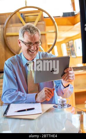 Zoom meeting in progress, mature businessman smiling in his barge boat office holding smart tablet Apple iPad computer in a virtual meeting situation Stock Photo