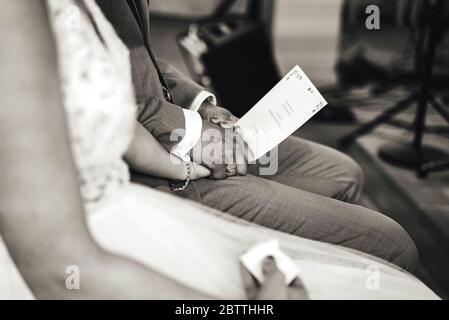 Black and white photo of sitting marriage couple at wedding ceremony. Close up of holding hands on grooms leg . Without head. Wedding day concept. Stock Photo
