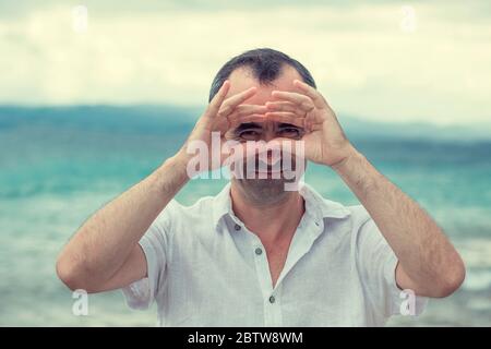 Closeup mature stunned curious man, peeking looking through fingers like binoculars searching something future forecast. Hipster male with beard while Stock Photo