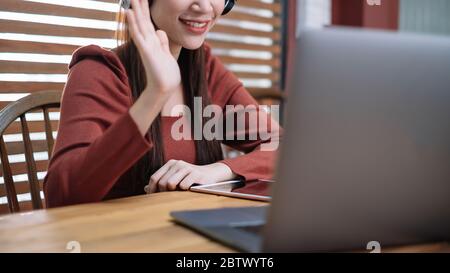 Smiling Asian businesswoman in headset waving hand, using laptop, looking at screen, student learning online Stock Photo