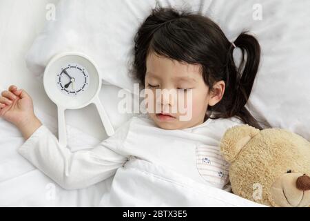 Child girl sleeping with teddy bear on the wooden bed in her bedroom, Happy asian child little girl and alarm clock wake up in the morning Stock Photo