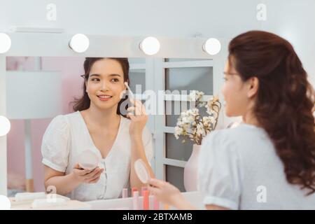 Portrait view of young adult asian woman looking at mirror, sitting in bright room and making make up. Beautiful and professional make up artist using Stock Photo