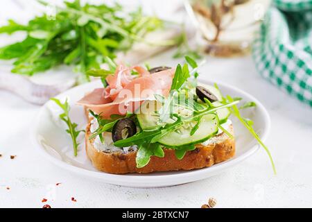 Sandwich with prosciutto, cucumber, black olives, arugula and feta cheese on  table.  Trend food. Stock Photo