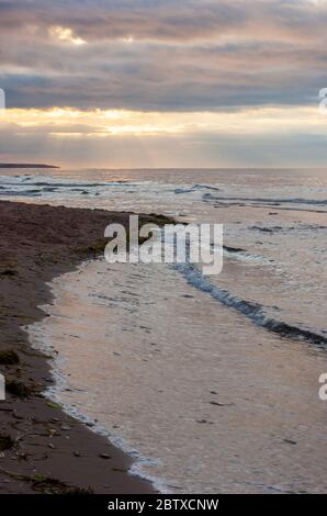 Dramatic sunset at Cavendish Beach, PEI National Park, Canada. Sunbeams shining through openings in clouds. Wavelets breaking on a deserted beach. Stock Photo