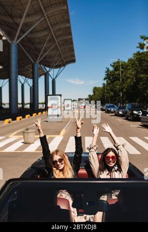 Two joyful girls in sunglasses happily looking in camera while driving cabriolet car on city streets outdoor Stock Photo
