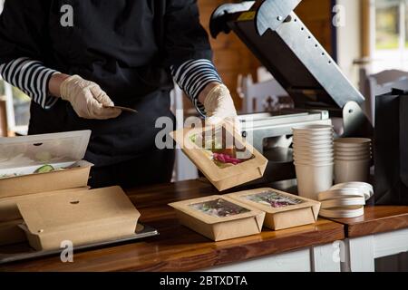 Restaurant worker wearing protective mask and gloves packing food boxed take away. Food delivery services and Online contactless food shopping. Stock Photo