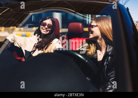 Joyful girls in sunglasses happily spending time together driving cabriolet on city streets outdoor Stock Photo