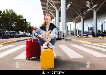 Pretty smiling girl in headphones happily using the tablet with suitcase near with airport on background Stock Photo