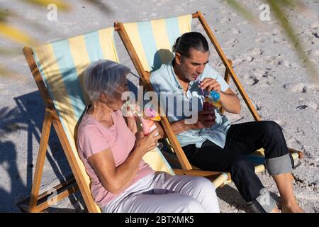 Senior Caucasian couple enjoying time at the beach on a sunny day, sitting on deck chairs, drinking drinks with sand in the background Stock Photo