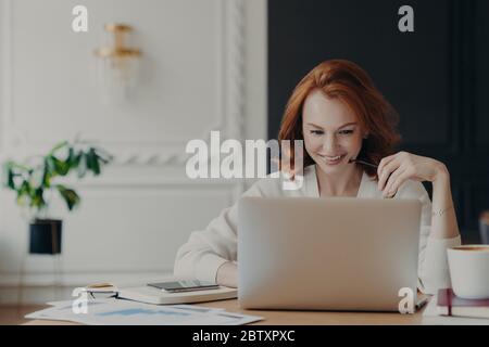 Positive happy female freelancer has busy working day, works distantly from home, sits in front of laptop computer against modern interior, works on c Stock Photo