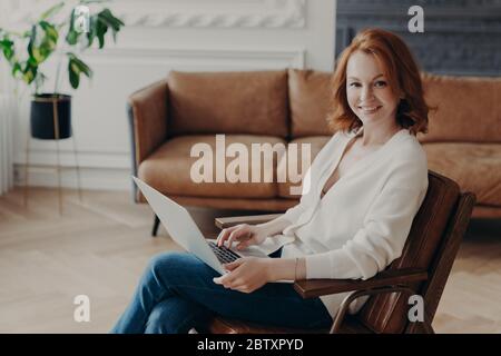 Sideways shot of woman entrepreneur sits in comfortable armchair in modern apartment with luxury furtniture, communicates with colleague through onlin Stock Photo