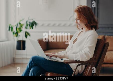 Smiling red haired woman freelancer sits in armchair, keyboards on laptop computer, uploads necessary material for developing site, poses in modern ap Stock Photo