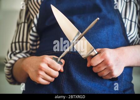 A cook in a blue apron and striped shirt is about to sharpen a kitchen knife on a long whetstone before cooking. Stock Photo