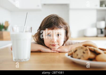 Cute toddler girl drinking milk with steel straw from glass and eating cookies sitting at kitchen table. Reduce plastic use at home with children Stock Photo
