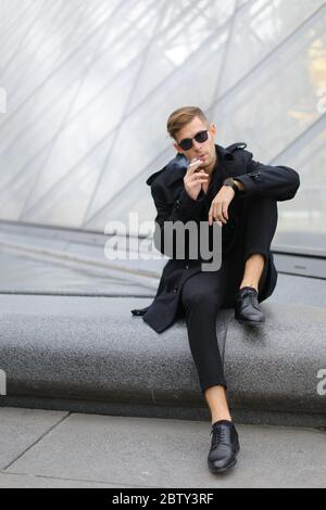 Young blong man wearing black jacket sitting on Louvre Pyramid and smoking. Stock Photo