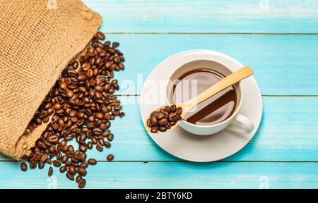 Cup of hot coffee and roasted coffee beans from the sack bag On a wooden table and spoon at the background of a blue wooden floor Stock Photo